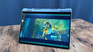 Dell 14 Plus 2-in-1 (Intel) laptop in tent mode on a wooden desk, against a black backdrop.