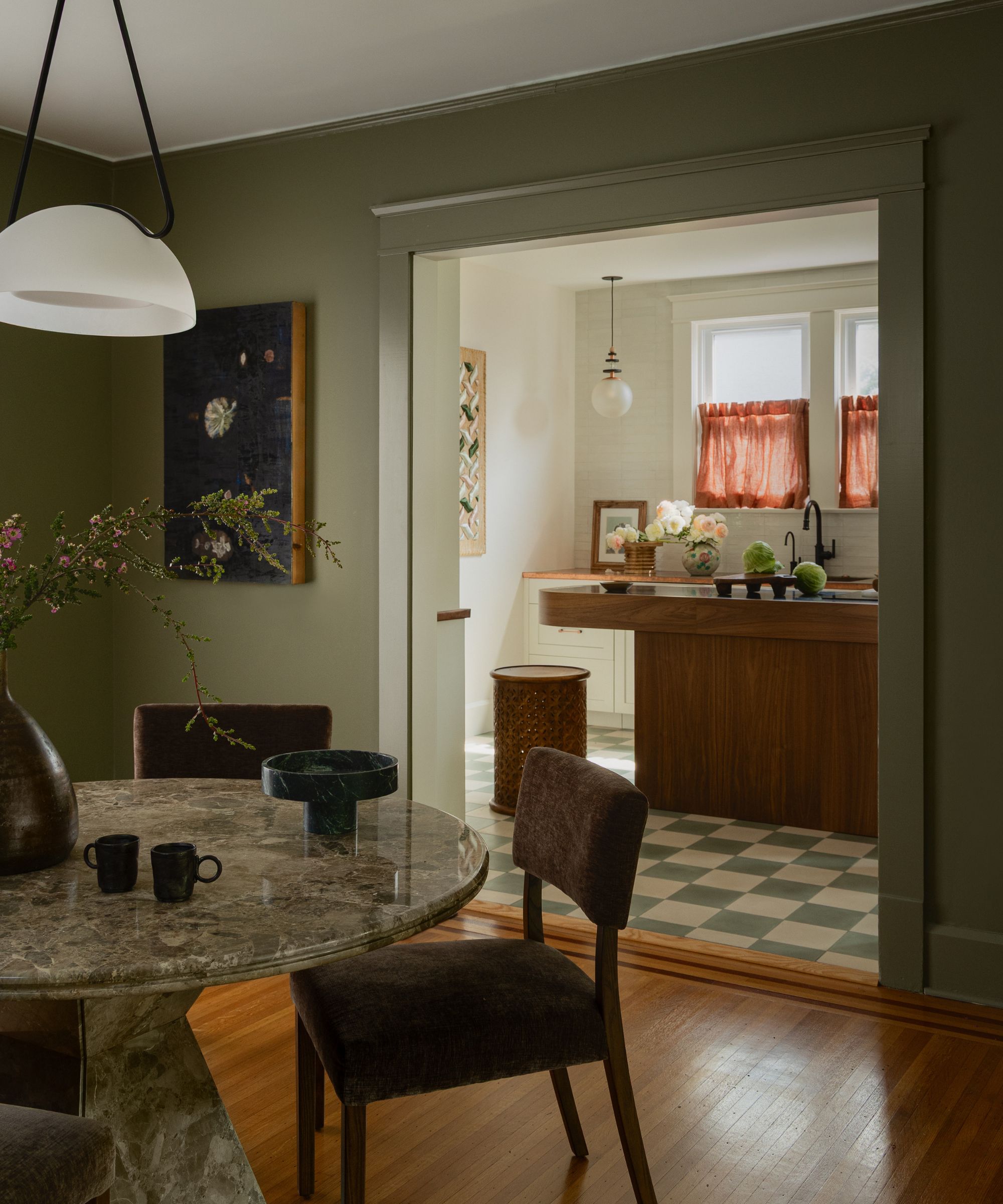 cozy kitchen with checkerboard floor, a wood island, orange cafe curtains with an open doorway through to the dining room painted an earthy green with a green marble round table