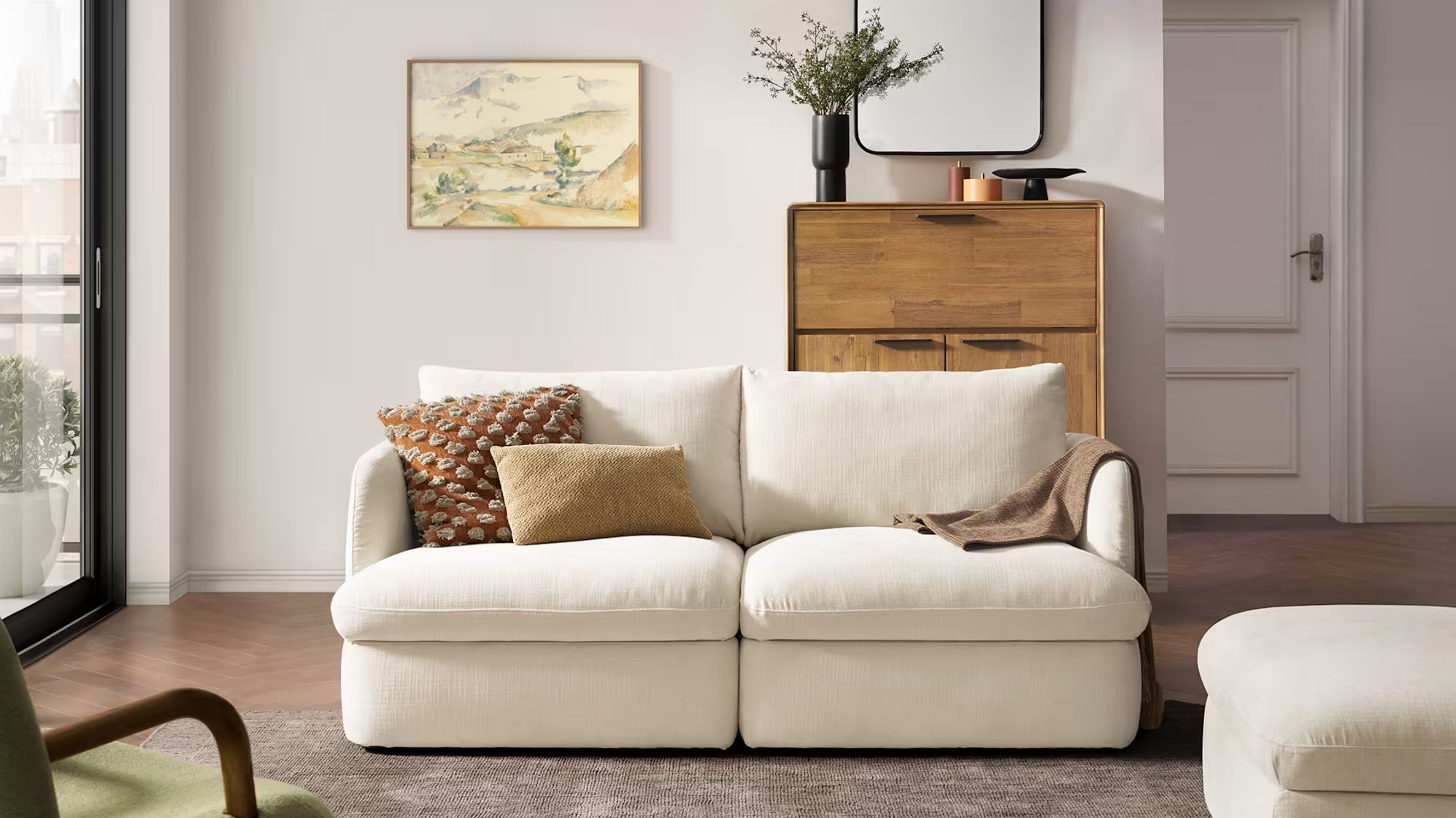 Image of a two-seater white sofa in a white living room with a gray rug underneath and a wooden console and painting on the back wall.