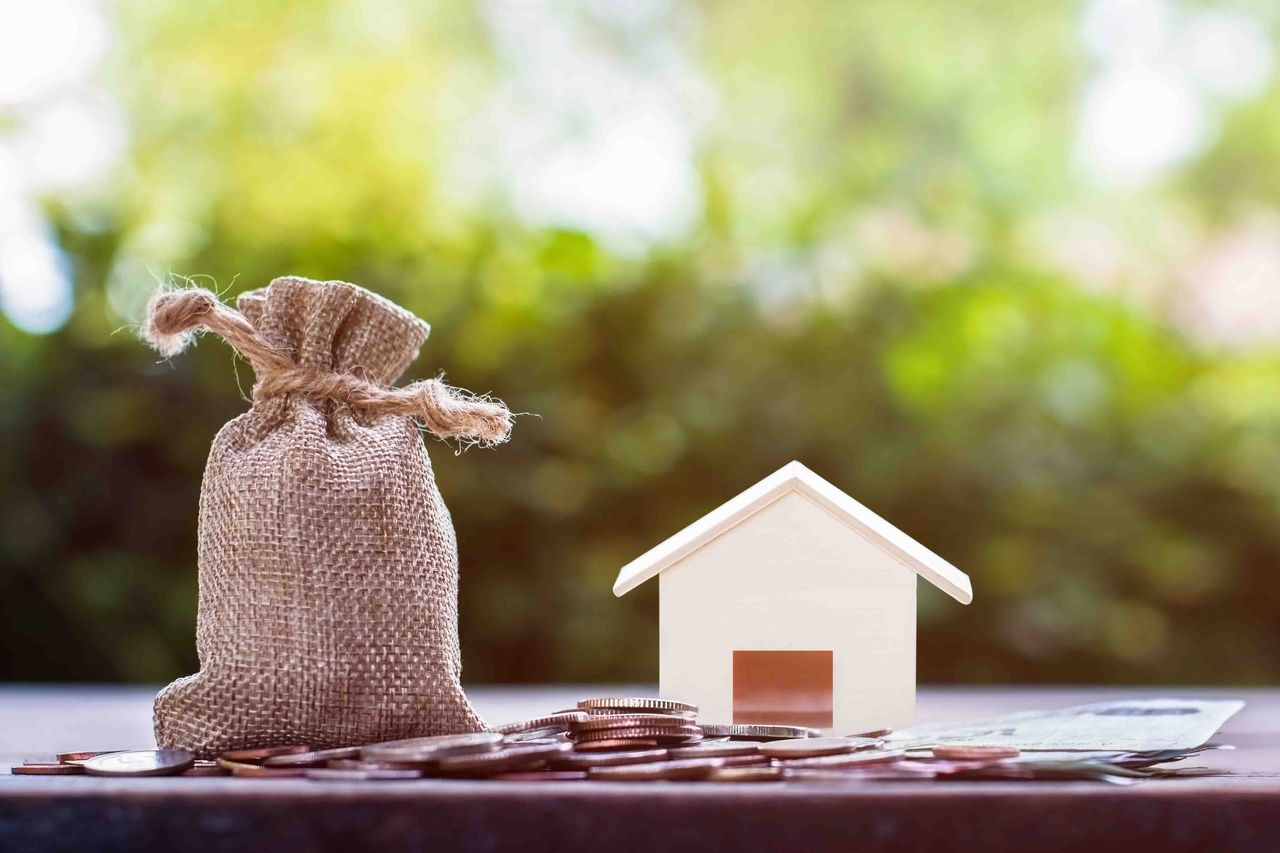 A bag of money next to a toy house with a pile of coins in front of it, representing a mortgage. 