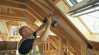 Steve who is wearing a hi-vis vest uses a drill to drill a hole in a wooden truss in the home's frame