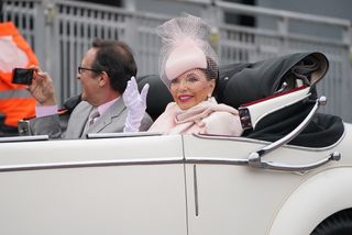 Joan Collins wearing a pink coat and fascinator riding in a white convertible car and waving during the 2022 Platinum Jubilee