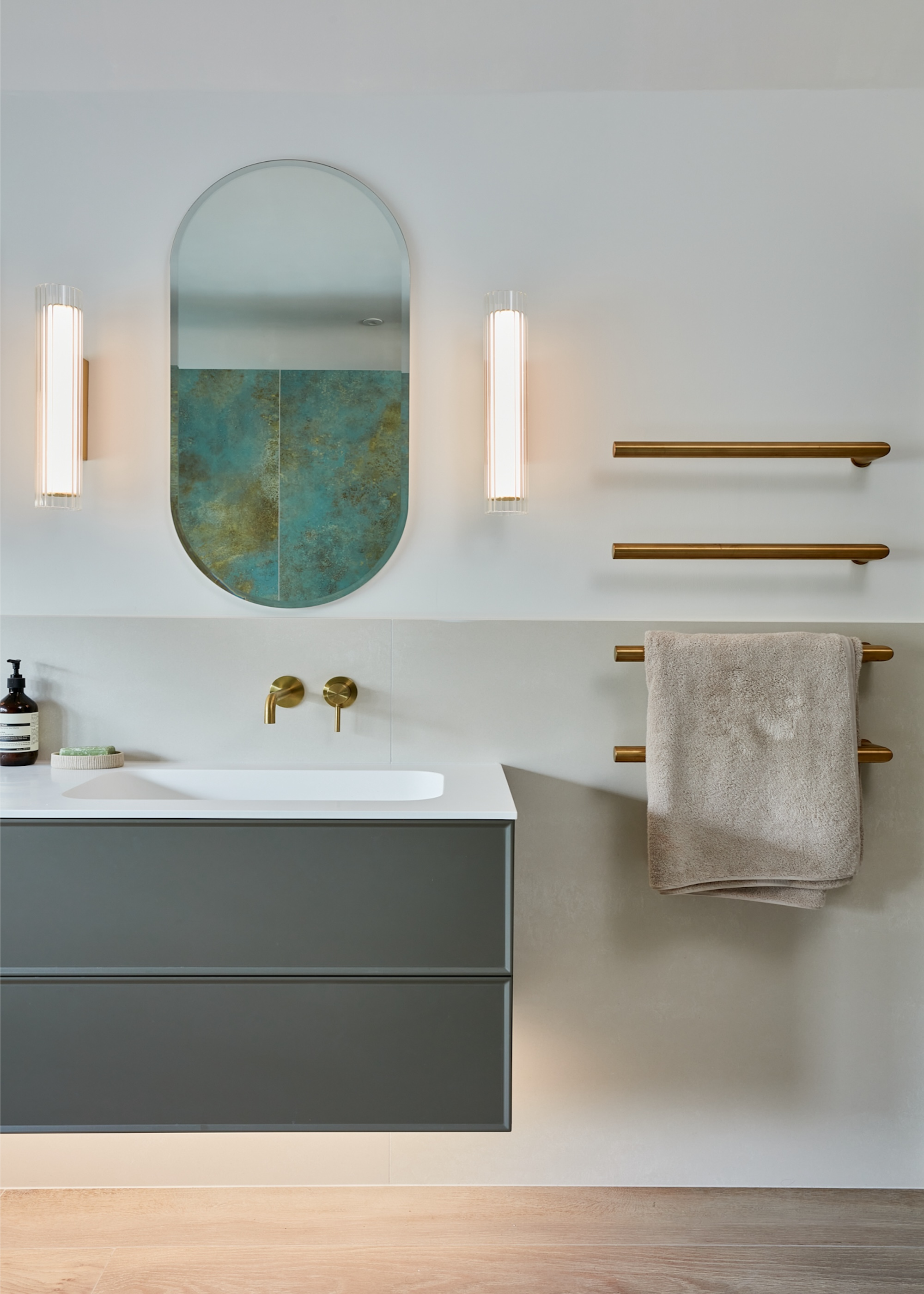 A bathroom with a floating vanity unit, gold towel rails and an oval-shaped mirror with wall sconces either side