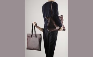 The torso and legs of the back of a woman wearing a black shirt, dark pants and holding a large leather handbag under one arm.
