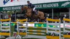 Ben Maher jumps a bay horse over a huge yellow and white triple bar jump in front of the Spruce Meadows crowd