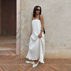 An image of an influencer wearing a strapless white dress, white crochet flats and black sunglasses