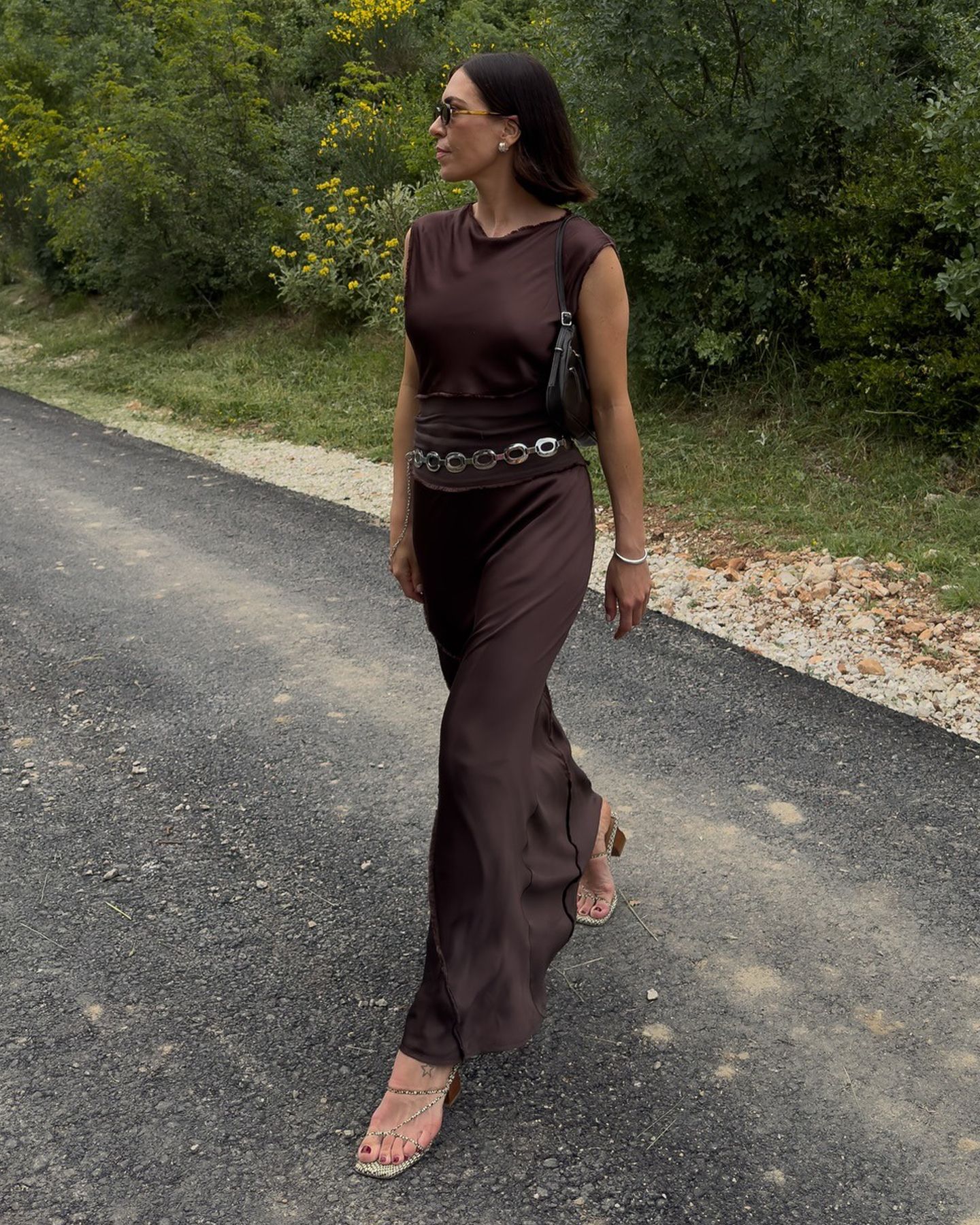 British style influencer Jessica Skye poses in a brown cowl-neck silk maxi dress with a silver chain belt, and strappy snakeskin sandals.