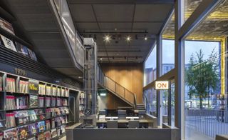 Interior view of the Hyundai Music Library featuring shelving units filled with records, a workspace with Apple Mac computers, an illuminated 'ON' sign, glass windows and stairs leading to the upper level