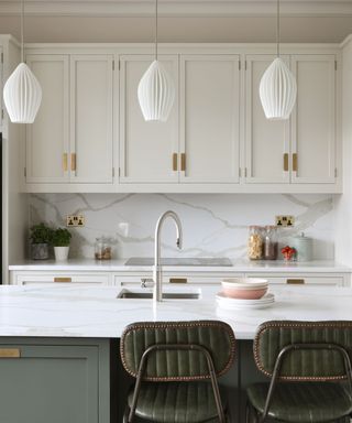 A marble kitchen island with a silver sink, sage green base, two green cushioned bar stools, three tulip pendant lights and white cabinets behind it