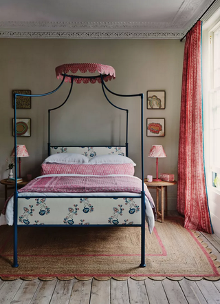 A taupe and red bedroom scheme with a jute rug, floral patterned headboard on a metal four poster bed and red patterned accents in the drapes, lighting and bed linen.
