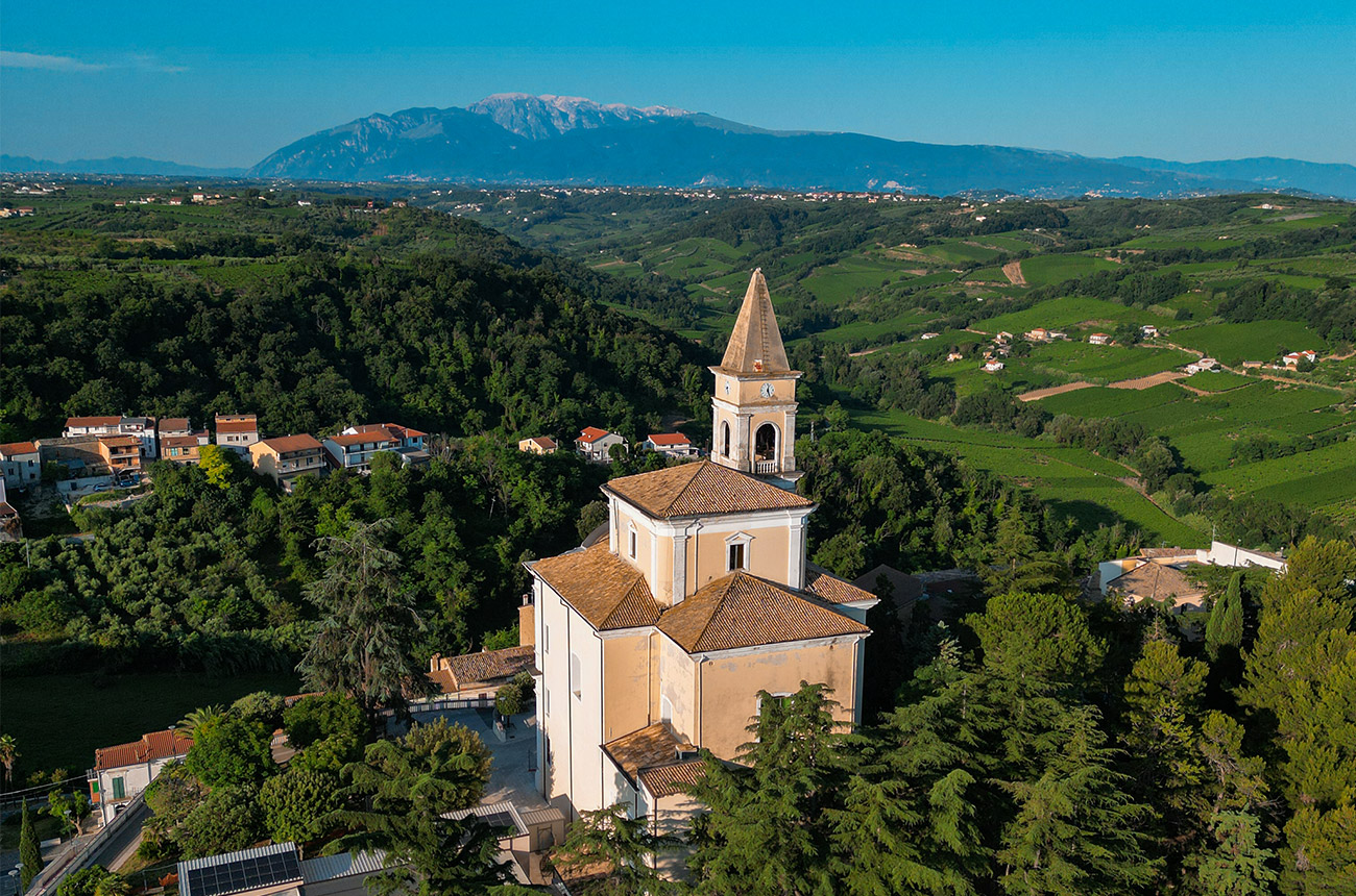 A large peach-coloured church surrounded by trees, small houses and green fields, with mountains in the background