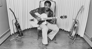 George Benson at home, in his element, pictured seated with his Ibanez, two guitars on stands on either side of him, in this archive B/W portrait from 1977.