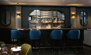 Interior view of the bar at Disgruntled Chef featuring grey walls, spotlights, two oval mirrors, blue chairs and a mirrored wall unit with bottles of drinks and glasses behind the bar
