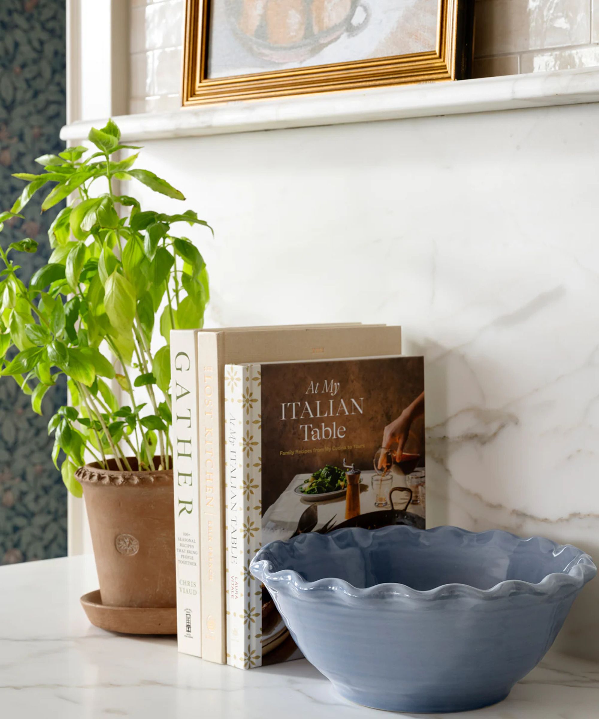 The corner of a marble kitchen displaying a terracotta plant pot, three recipe books, and a blue bowl with ruffled edges