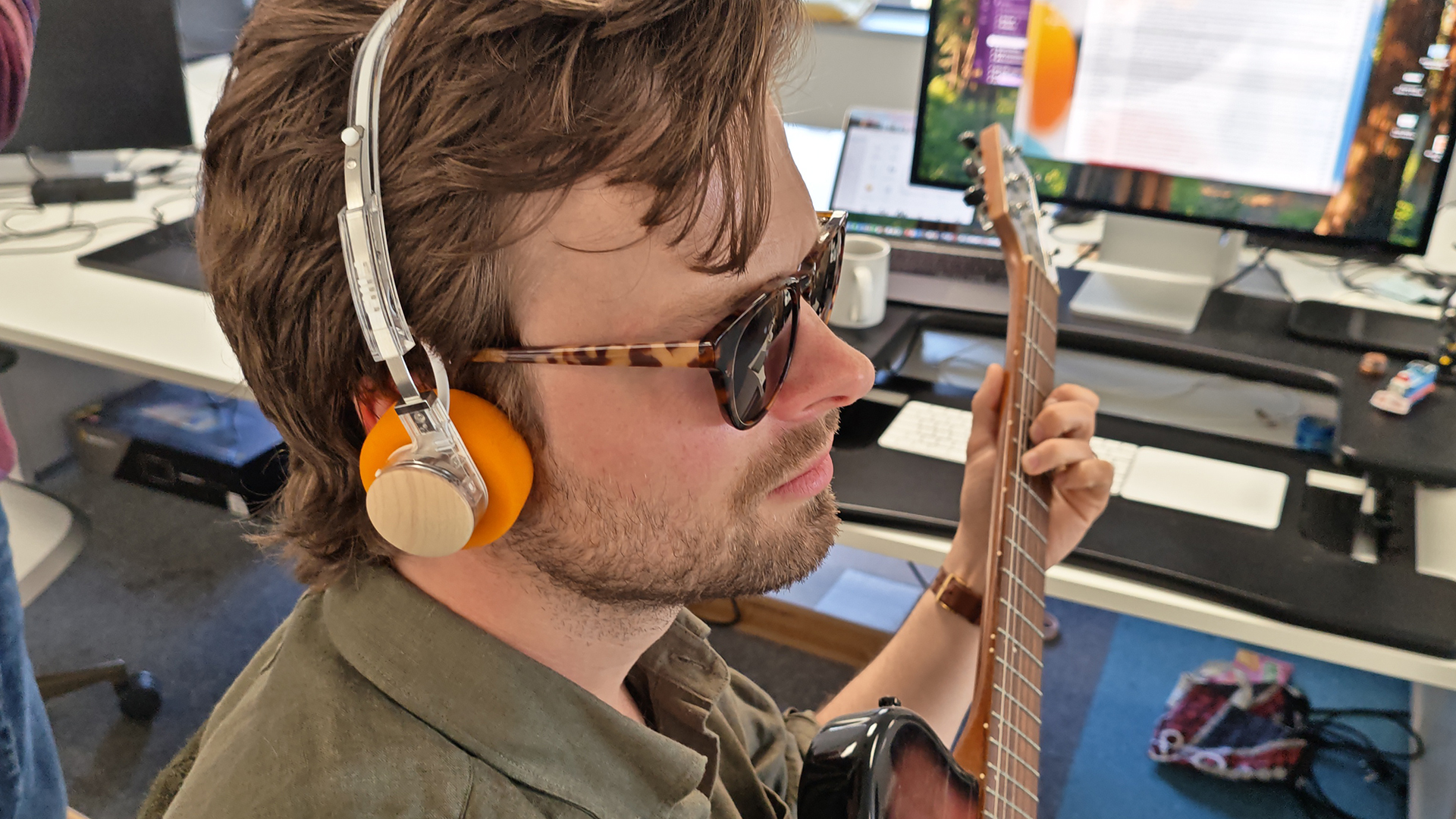 A man wearing orange headphones, plays a brown guitar, in an office. 