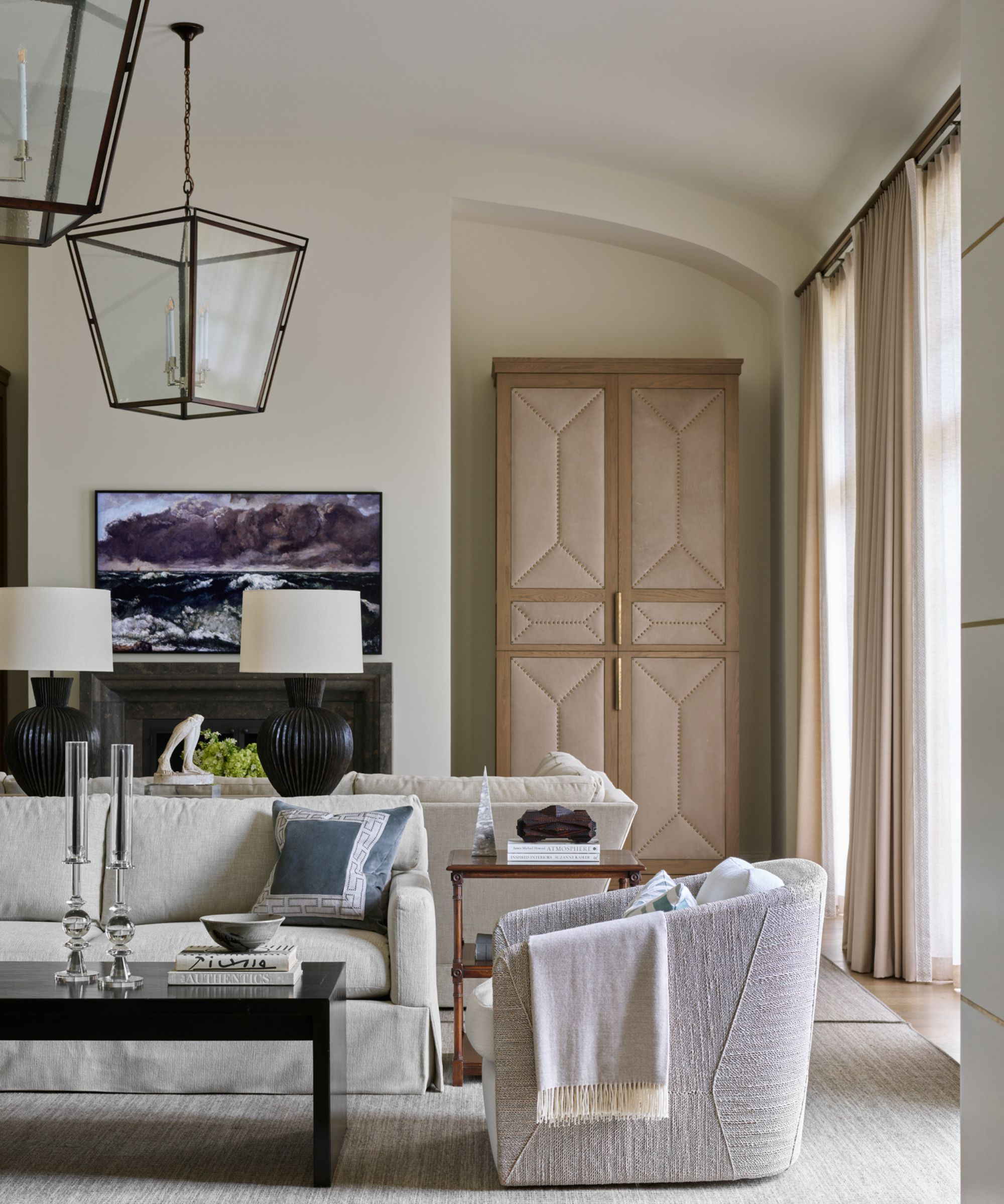 Large living room with high ceilings, lantern glass fixtures, cream sectional sofa, a curved boucle armchair, large windows with cream drapes, and a black coffee table with candlesticks on top.