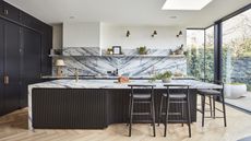 A dark blue black kitchen showing an island with fluted kitchen detail, marble worktops and splashback and gold taps and hardware
