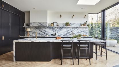 A dark blue black kitchen showing an island with fluted kitchen detail, marble worktops and splashback and gold taps and hardware