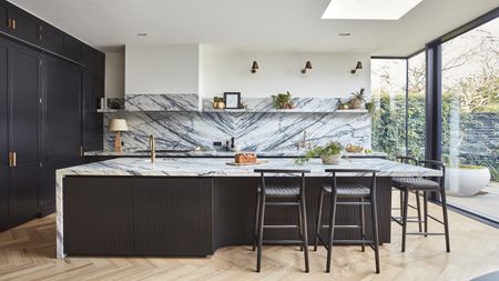 A dark blue black kitchen showing an island with fluted kitchen detail, marble worktops and splashback and gold taps and hardware