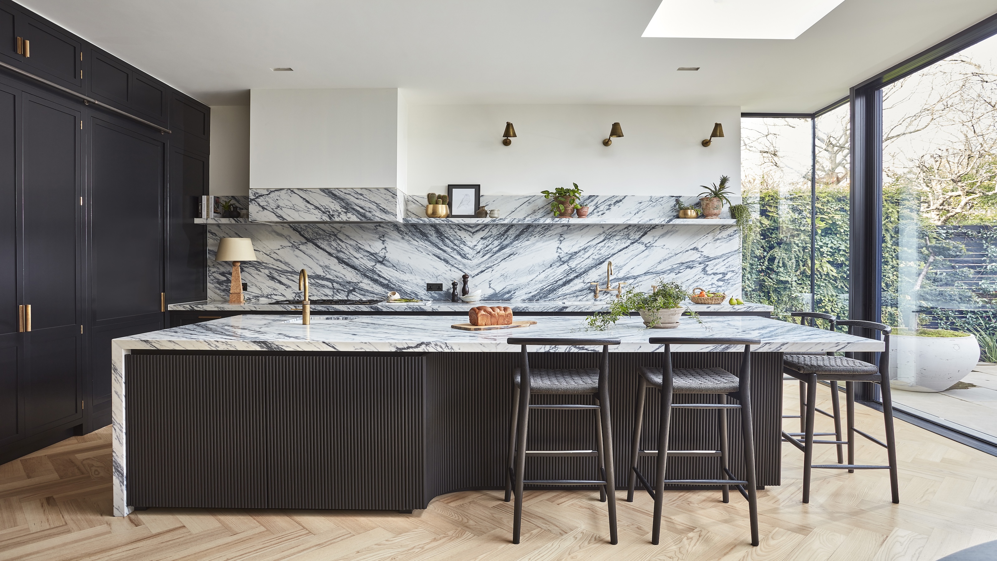 A dark blue black kitchen showing an island with fluted kitchen detail, marble worktops and splashback and gold taps and hardware