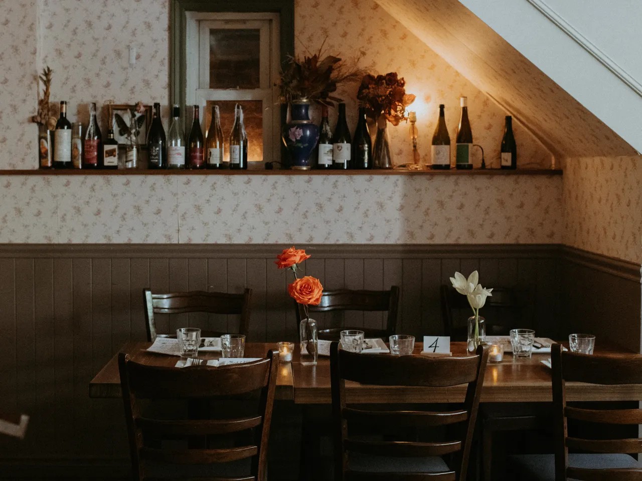 A dimly lit restaurant with shabby chic furniture and interiors, including wood-paneled walls and wallpaper with floral designs, wooden furniture, and a collection of bottles overlooking a table.