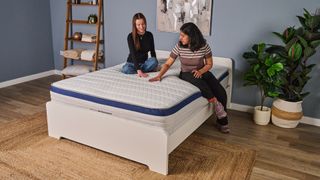The image shows two women sitting on the updated Helix Midnight mattress, which is placed on a white bed frame in a neutrally decorated bedroom