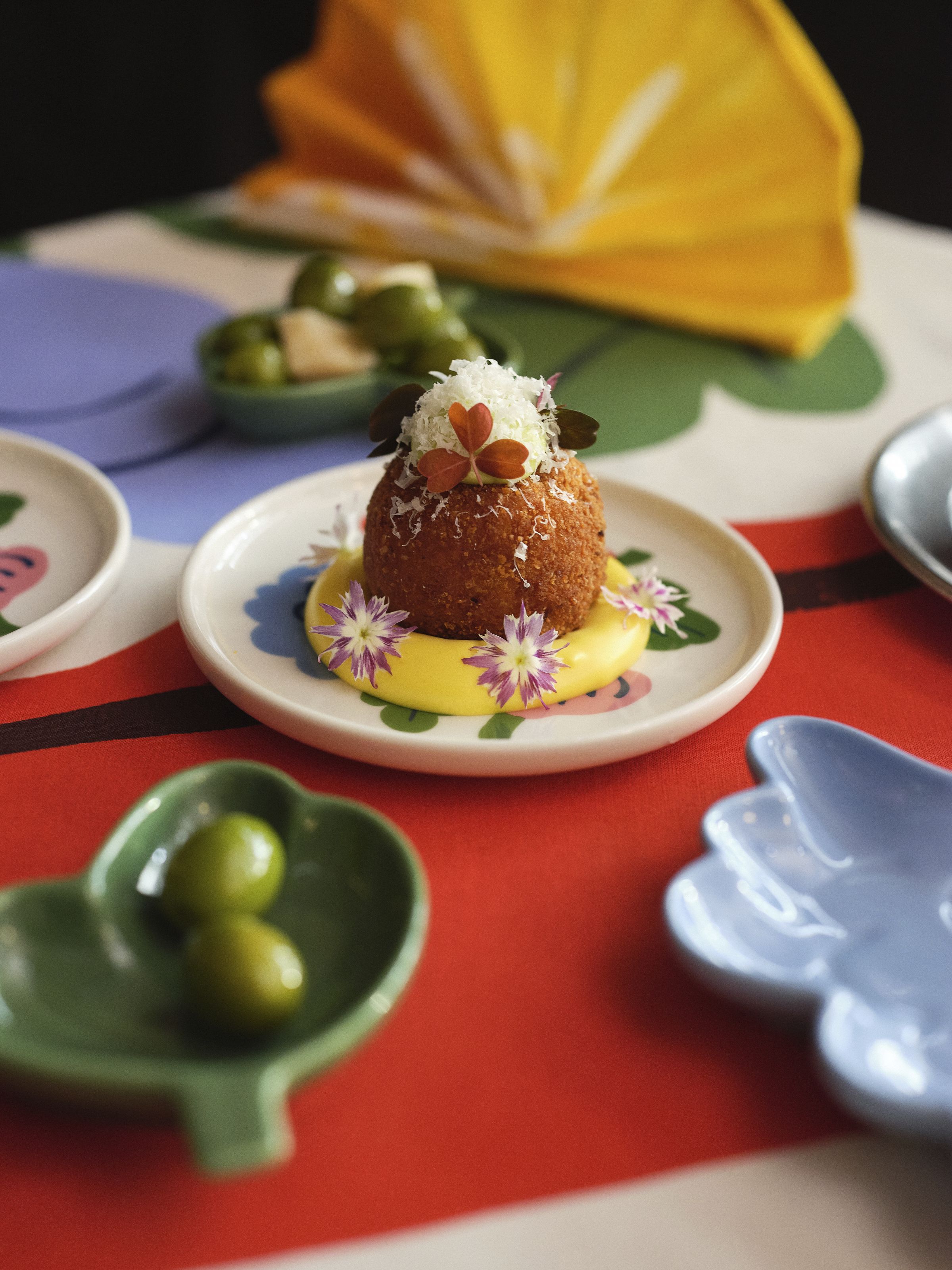 The exterior of an osteria, decorated with a floral mural, a table topped by floral-shaped serveware, and a woman dressed in a gray jumper and a white, brown, and blue skirt, handling yellow tuplis.
