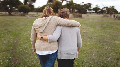 An adult woman and her mother walk with arms around each other outside.