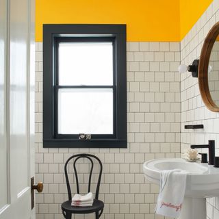 budget bathroom ideas, white square tiles with top quarter of walls painted bright yellow, window frame painted black, black fixtures, black chair, towel on sink