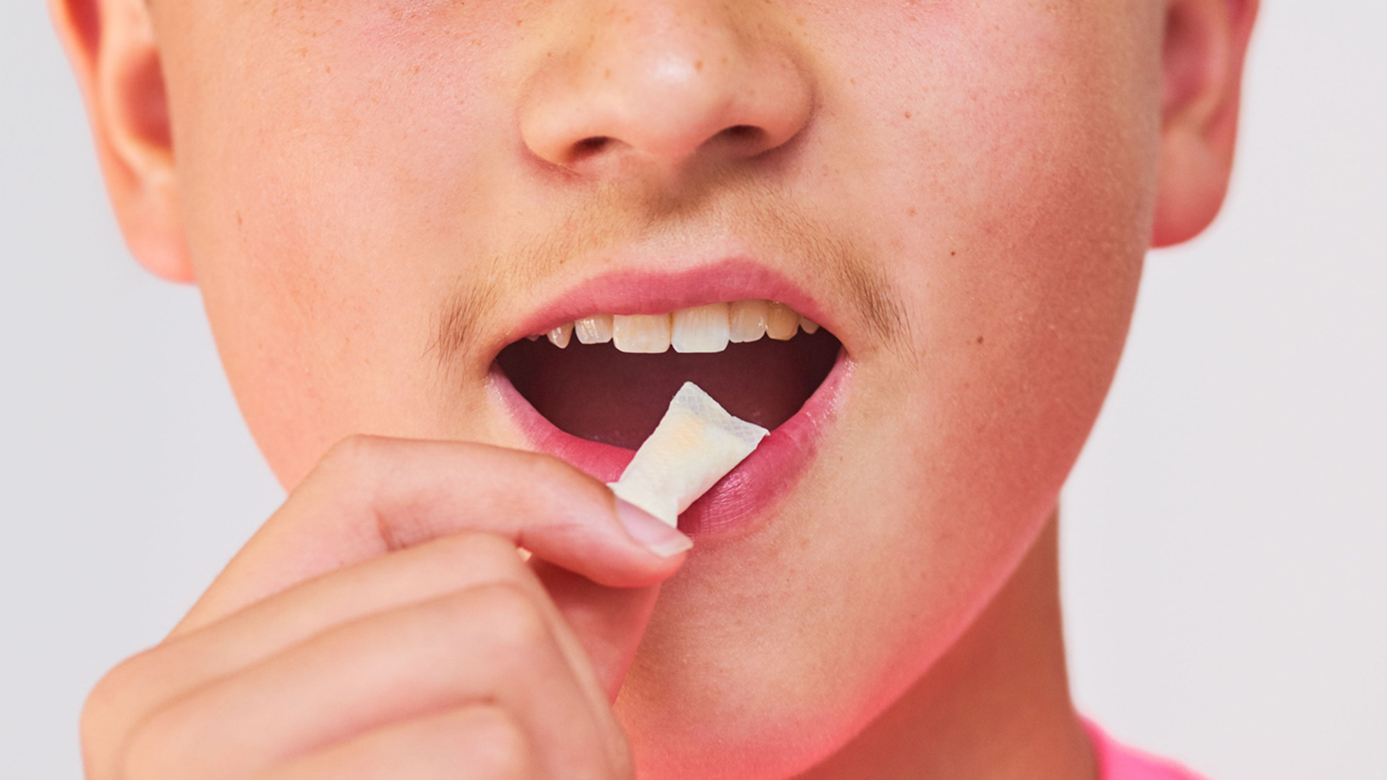 A close up of a male teenager's mouth where he holds a small rectangular object in his open mouth