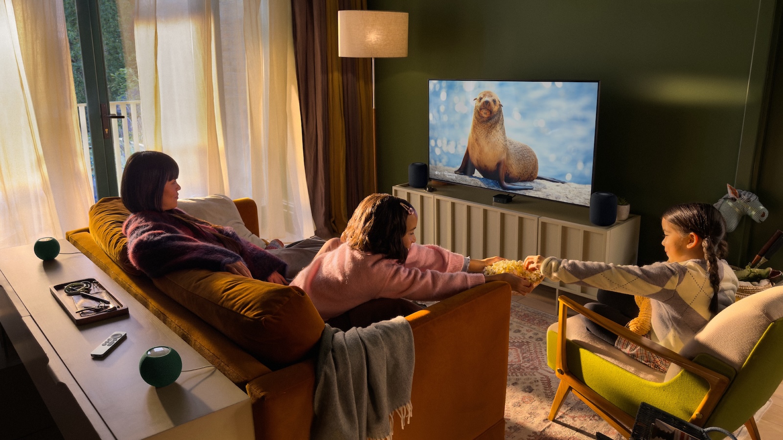A photograph of a family watching TV, with four HomePod speakers dotted around the room for sound