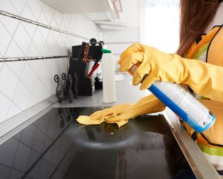 Woman with long brown hair wearing yellow gloves, spraying and cleaning a black electric stove top