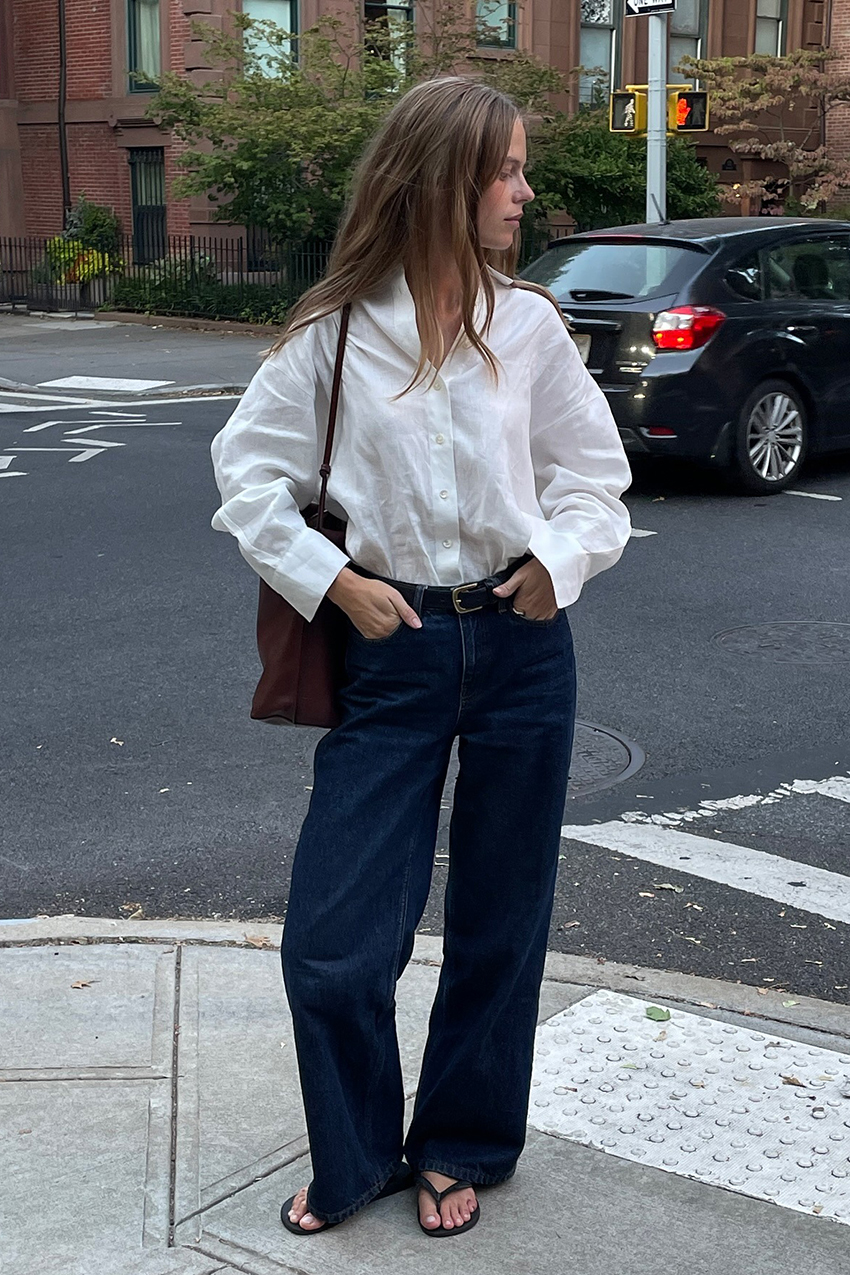 a woman wearing a white button-down, denim blue jeans, flip flops, and brown leather bag