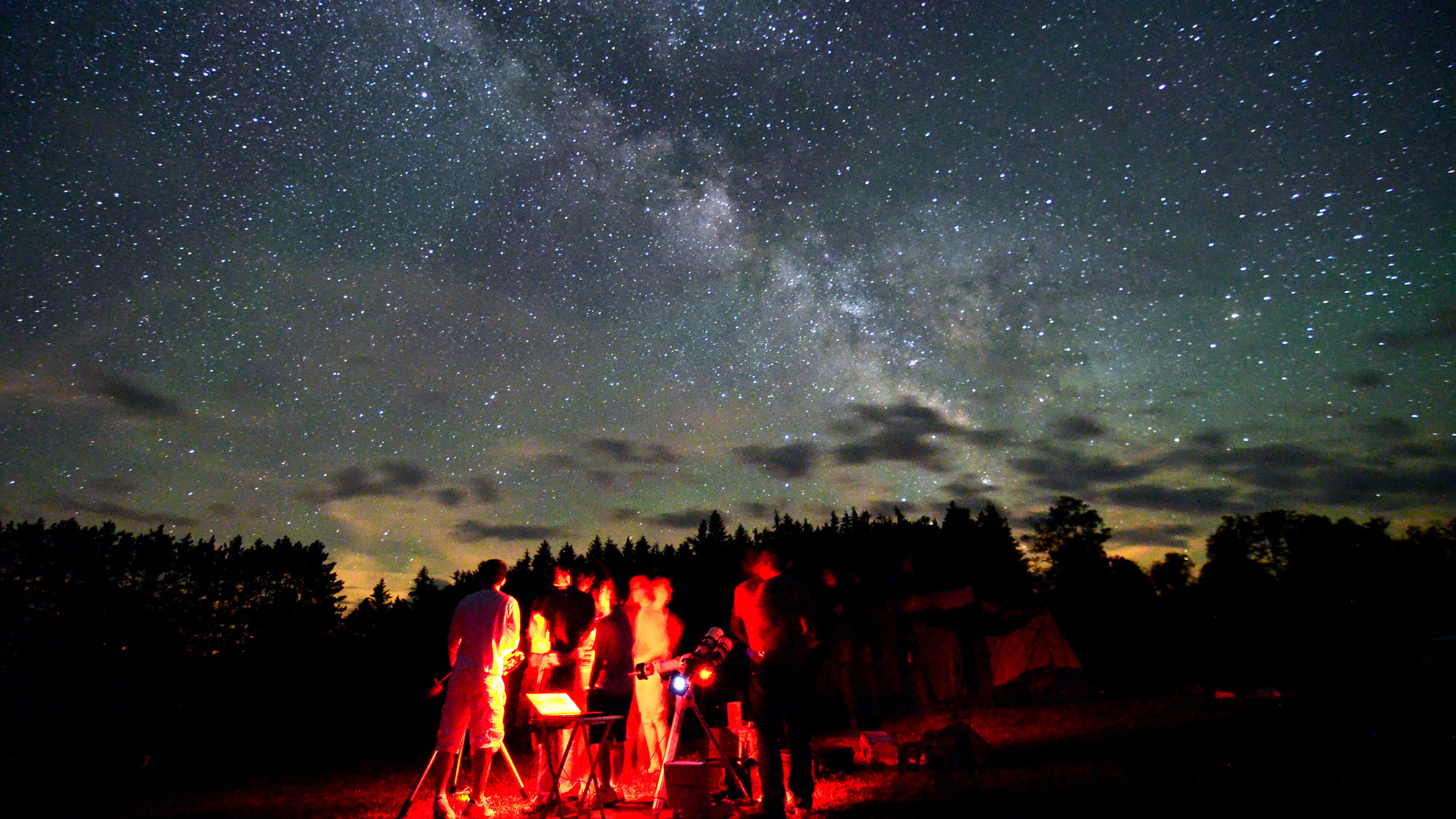 A group of people stand under a red light and look up at a deep blue night sky.