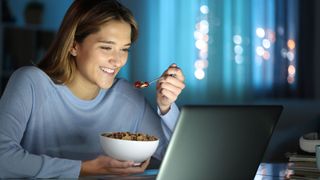 A blonde woman eats cereal at night while looking at laptop screen