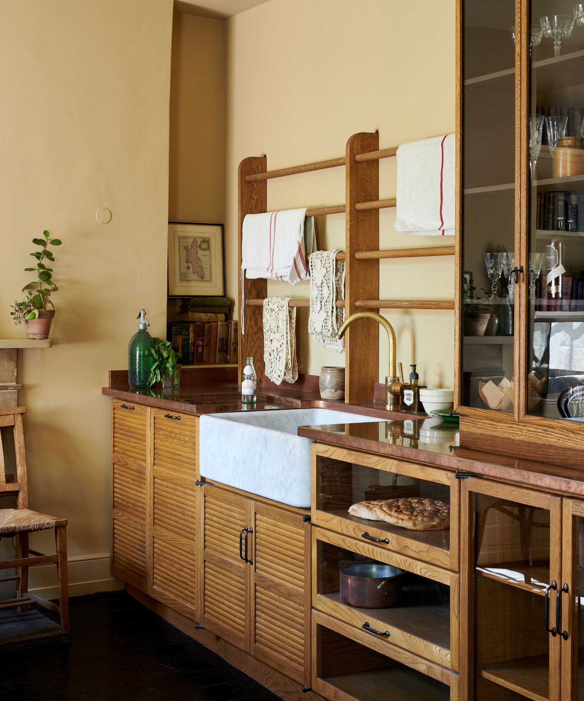 Warm kitchen with yellow walls, wooden slatted cabinets, a white marble farmhouse sink with a brass faucet, and a wall-mounted wooden towel drying rack
