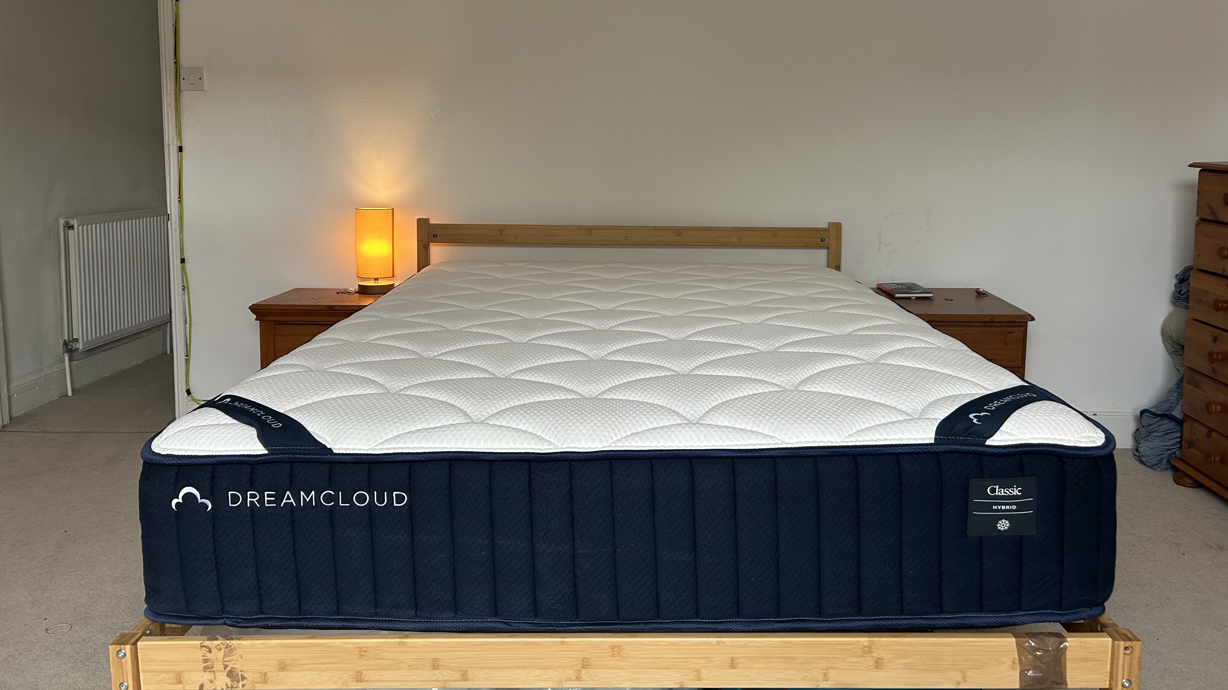 The DreamCloud Classic Hybrid mattress on a wooden bedframe with two wooden bedside tables beside it a small orange lamp lit up