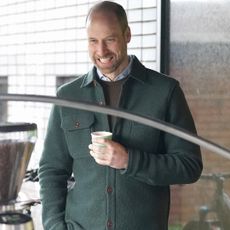 Prince William wearing a green shacket holding a coffee cup and smiling 