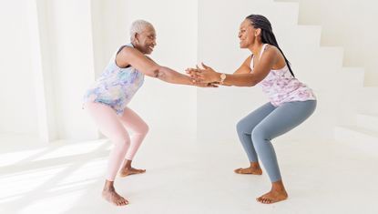 Two women squat while facing each other. They are in a sparse, white space, and are wearing athletic leggings and tank tops, and are barefoot 