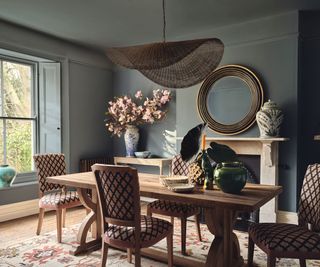 blue dining room with wooden dining table and upholstered dining chairs and round mirror with statement wicker pendant light