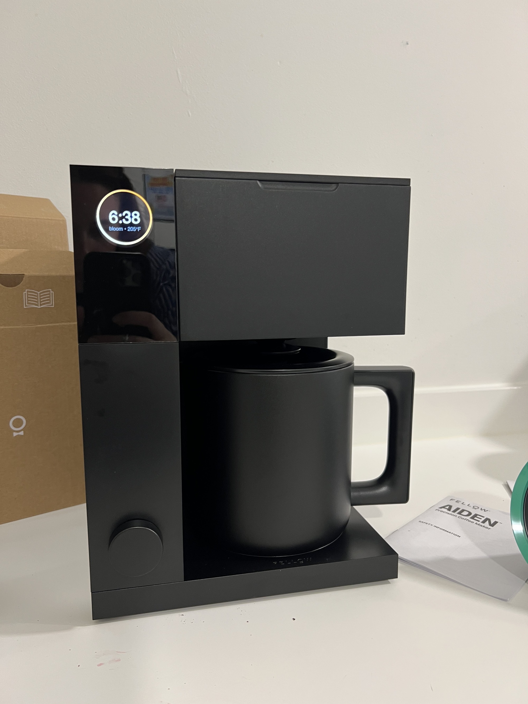 A black Fellow Aiden Precision coffee maker on a white countertop with a cardboard box behind it.