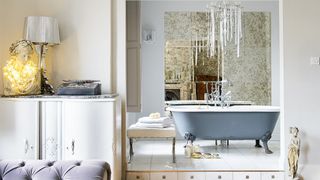 luxurious en-suite bathroom in a luxurious home, with a grey roll-top bath, glittering tiles, a chandelier, and a white chest of drawers and purple chaise lounge in front