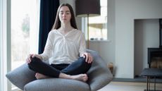 woman wearing a white shirt and dark trousers sitting on a single sofa chair with a living room setting behind her. 