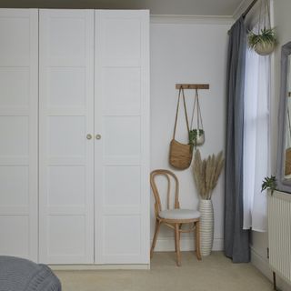 White cupboard next to a bentwood chair an a white wall with an accessory rack , a vase with décor and a grey curtain