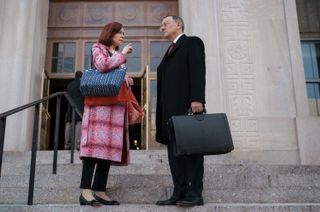 carrie preston as esbeth wearing a pink plaid coat talking to michael emerson in a suit as Judge Milton Crawford outside of a court house