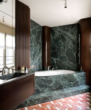 Green marble bathroom with fluted wood paneling and earthy pink floor tile surrounding corner tub