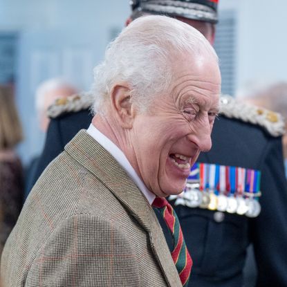 King Charles wearing a tan blazer and striped tie turning to the side and laughing surrounded by a group of people 