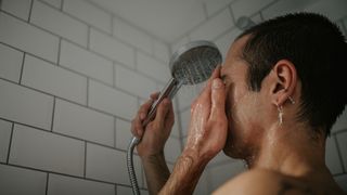 A man showers in a white, tiled bathroom, focusing on his face