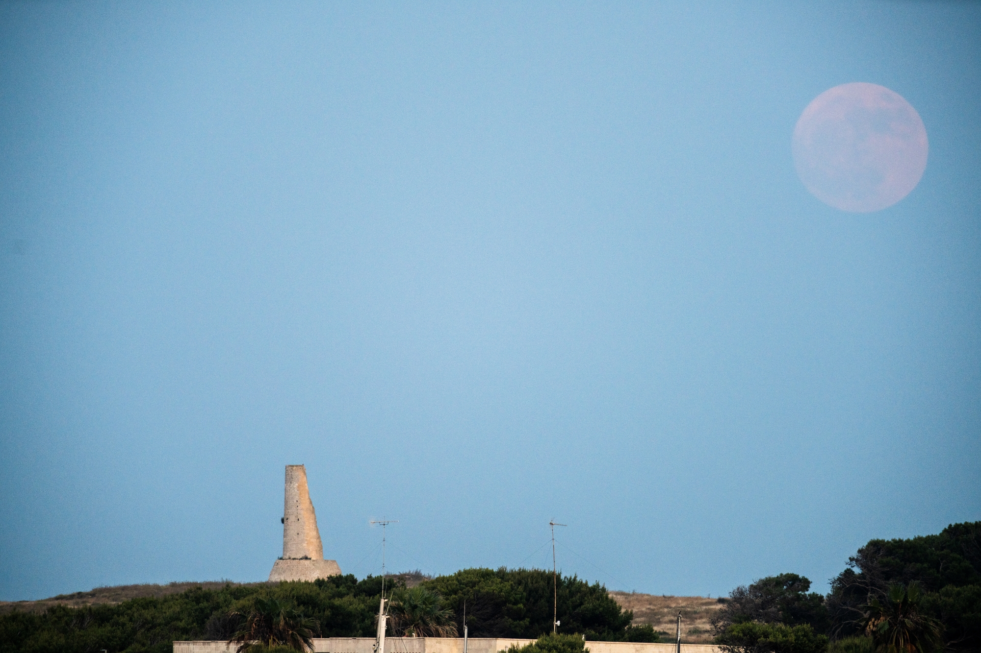 A photograph of the Strawberry Moon rising over the Torre del Serpe in Otranto, Italy.
