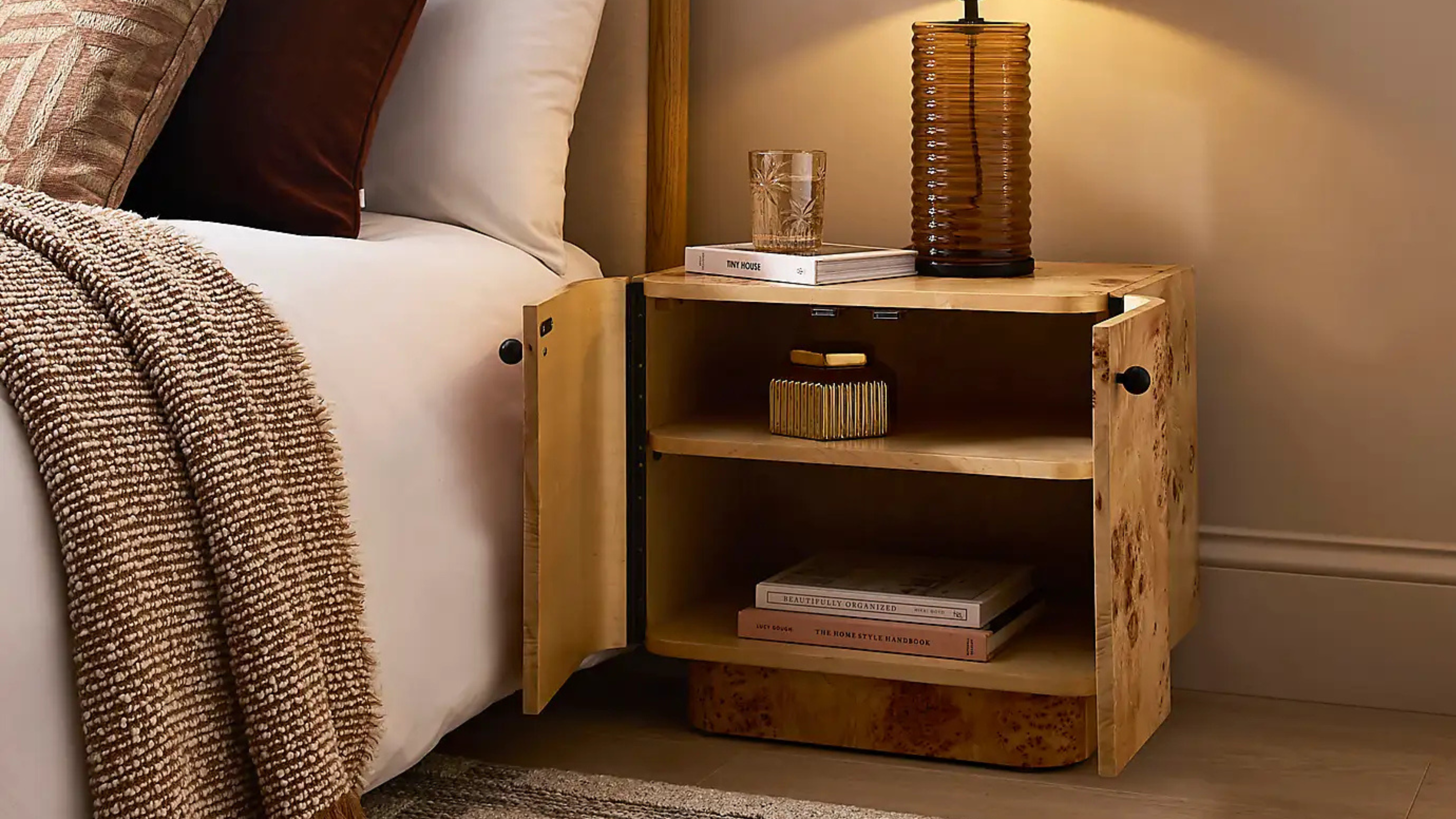 dunelm&#039;s seana burl bedside table with it&#039;s two doors open with books and a storage container inside with a lit lamp on top of the bedside next to a styled bed